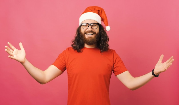 Foto de homem alegre usando chapéu vermelho de papai noel mostrando gesto de boas-vindas sobre fundo rosa