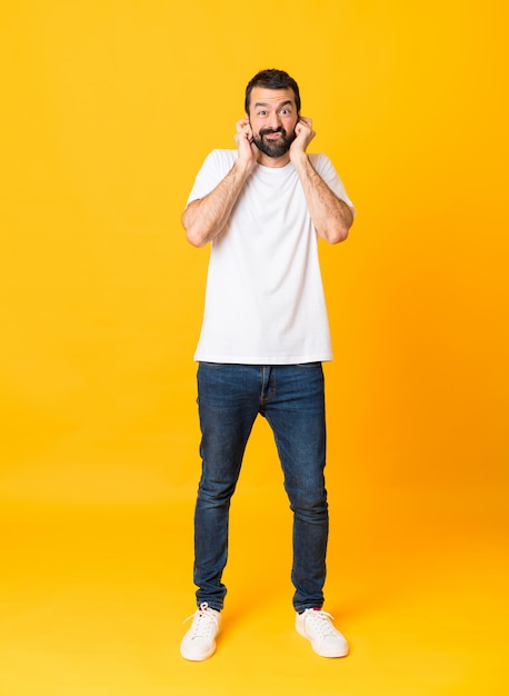 Foto de homem com barba sobre amarelo isolado frustrado e cobrindo as orelhas