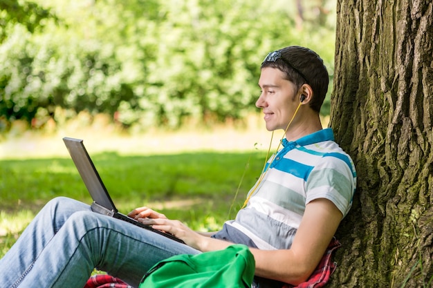 Foto de homem com laptop e fones de ouvido sentado sob uma árvore no gramado de verão