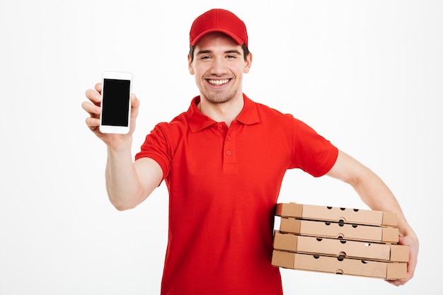 Foto de homem feliz do serviço de entrega em t-shirt vermelha e boné segurando a pilha de caixas de pizza e mostrando o smartphone, isolado sobre o espaço em branco