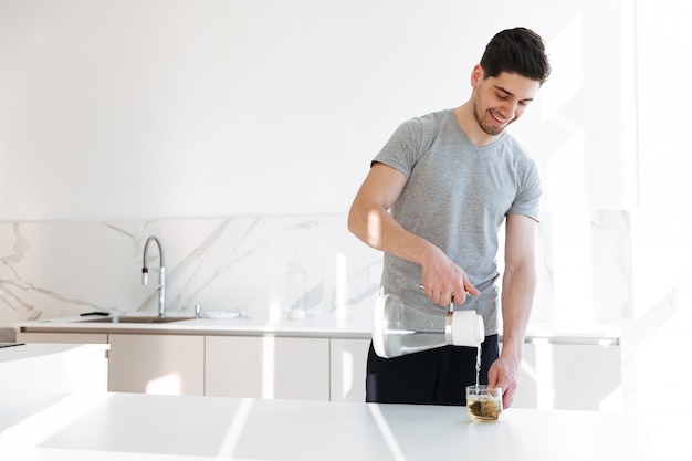 Foto de homem musculoso em camiseta casual, fazer chá com despejar água quente na xícara, enquanto tomando café da manhã na manhã em casa