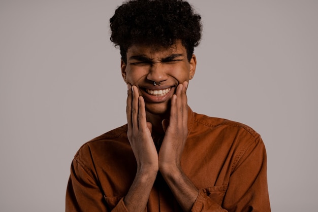 Foto de homem negro parece ter dor nos dentes. Homem usa camisa, fundo de cor cinza isolado.