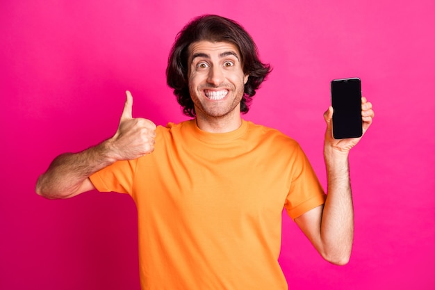 Foto de homem segurando telefone espaço vazio levantar polegar para cima sorriso branco usar camiseta laranja isolado fundo de cor rosa