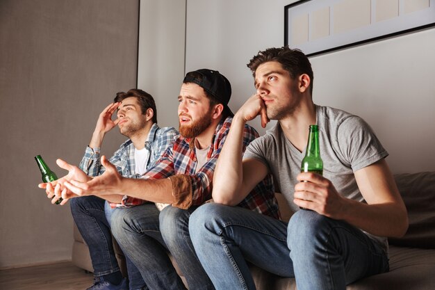 Foto de homens adultos descontentes expressando frustração, sentados no sofá e assistindo a um jogo de futebol no apartamento