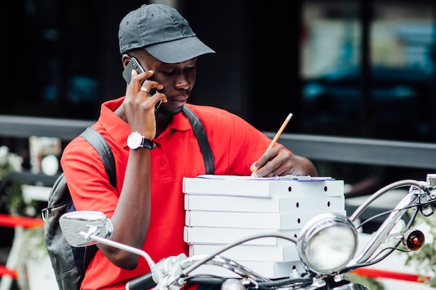 Foto de jovem africano aceita o pedido por telefone e escreve na motocicleta segurando caixas com pizza. lugar urbano.