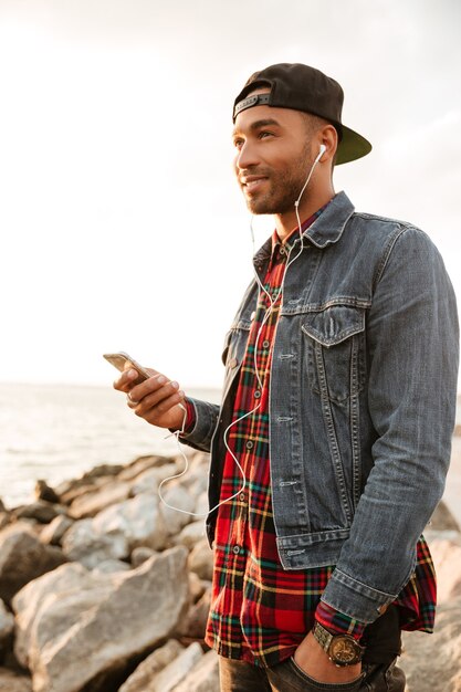 Foto de jovem alegre de boné, caminhando na praia e conversando ao lado do telefone enquanto ouve música