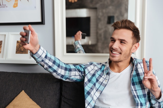 Foto de jovem alegre, vestido com uma camisa com uma impressão da gaiola, sentado no sofá em casa e tirar uma selfie.