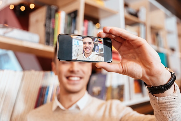 Foto de jovem alegre vestido com uma camisa, sentado no café e fazer selfie por seu telefone.