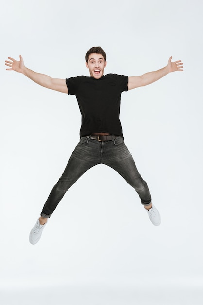 Foto de jovem alegre vestido com uma camiseta preta, pulando sobre um fundo branco, olhando para a câmera.