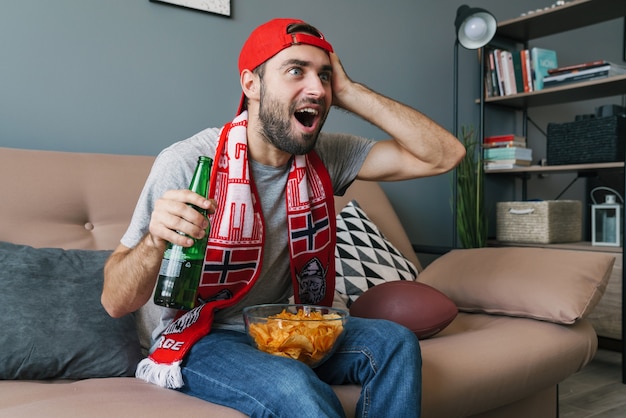 Foto de jovem animado com uma bola de rugby comendo batatinhas e bebendo cerveja enquanto assiste a uma partida de esportes na sala de estar