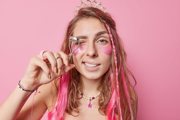 Foto de jovem cuida de sua aparência quer ficar bonita usa curvador de cílios aplica adesivos de colágeno sob os olhos faz maquiagem diária tem poses de cabelo comprido em ambientes fechados contra parede rosa