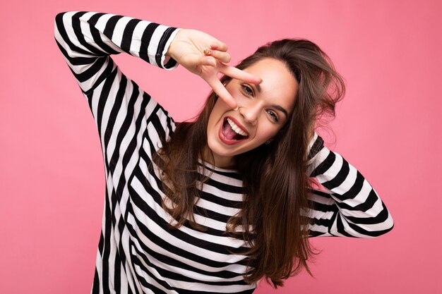 Foto de jovem europeu positivo sorridente atraente feliz linda morena feminina com emoções sinceras, vestindo pulôver listrado casual isolado no fundo rosa com espaço de cópia.