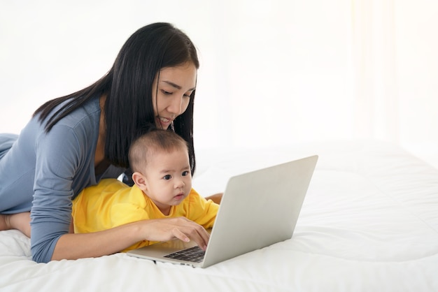 Foto de jovem feliz mãe tailandesa com seu bebê usando o laptop na cama. Conforto do lar. Cuidado e atenção. Trabalho a partir de casa.