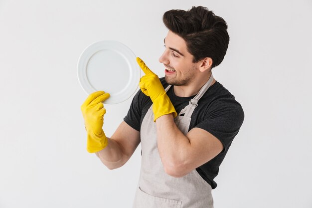 Foto de jovem feliz usando luvas de borracha amarelas para proteção das mãos, lavando pratos enquanto limpa a casa isolada sobre o branco