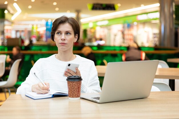 Foto de jovem morena atraente sentada em um shopping center em uma mesa com um copo de papel de café e trabalhando em um laptop de computador, usando o celular. Conceito de freelance e negócios.