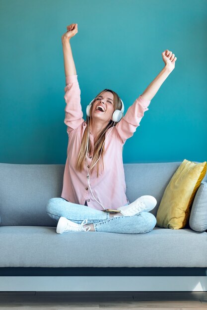 Foto de jovem motivada ouvindo música com o smartphone enquanto está sentado no sofá em casa.