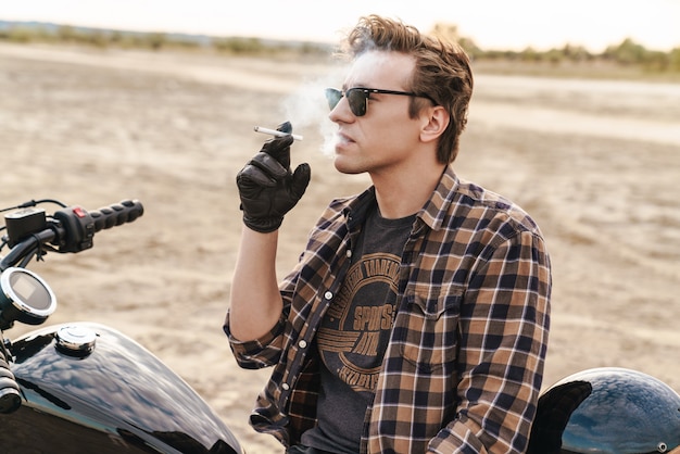 Foto de jovem motociclista ao ar livre no cigarro de fumo de campo no deserto.