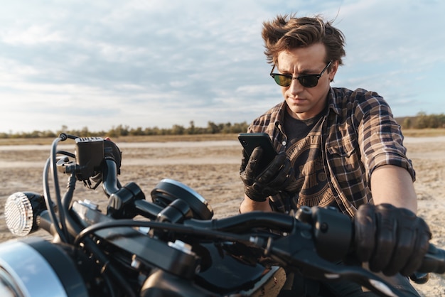 Foto de jovem motociclista em bicicleta ao ar livre no campo do deserto usando telefone celular.