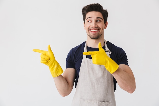 Foto de jovem otimista usando luvas de borracha amarelas para proteção das mãos apontando os dedos para copyspace enquanto limpa uma casa isolada sobre o branco