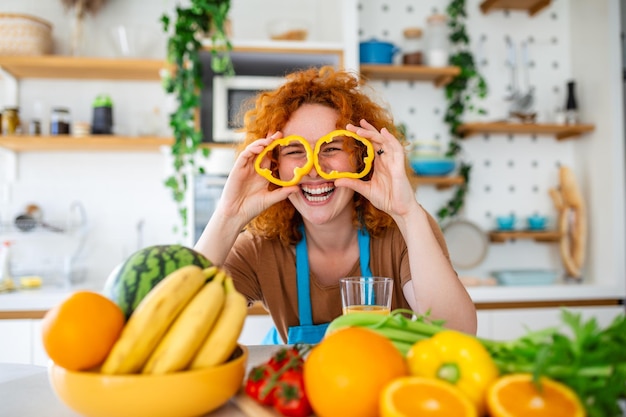 Foto de jovem sorrindo e segurando círculos de pimenta nos olhos enquanto cozinha salada com legumes frescos no interior da cozinha em casa