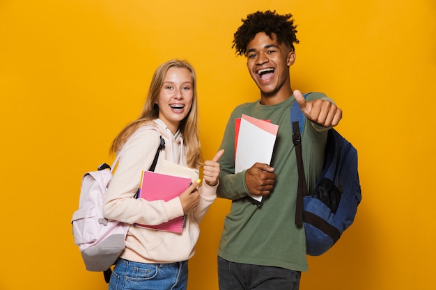Foto de jovens estudantes rapaz e garota de 16 a 18 anos vestindo mochilas, sorrindo e segurando cadernos, isolados sobre fundo amarelo