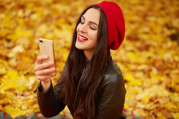 Foto de linda garota vestindo boina vermelha e jaqueta de couro preta no parque outono, fazendo o auto-retrato em seu celular