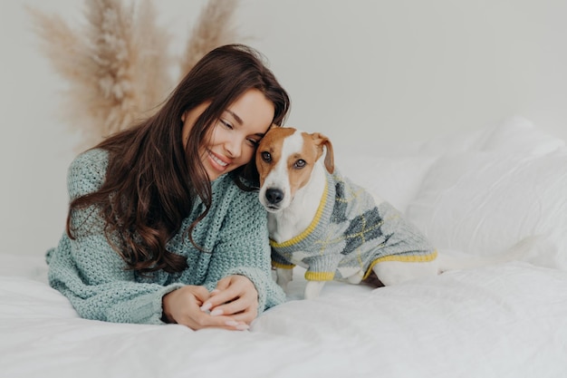 Foto de linda mulher morena de suéter de malha deitada junto com cachorro na cama macia gosta de passar tempo com o animal de estimação favorito se preocupa com animais ficar em casa durante a quarentena de coronavírus