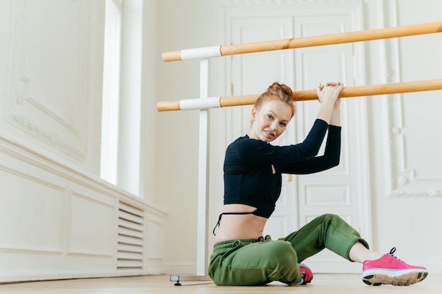 Foto de linda mulher ruiva faz exercícios de pilates no corrimão senta-se no chão se estica no estúdio de dança estando em boa forma corporal vestida com roupas esportivas Esporte e cuidados de saúde