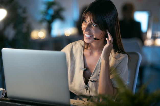 Foto de linda sorridente jovem mulher de negócios trabalhando com o laptop enquanto fala com fone de ouvido, sentado no escritório.