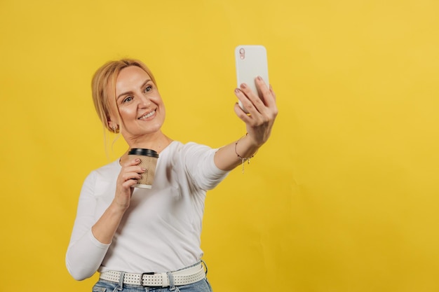 Foto de loira madura e fofa tira selfie usando camiseta branca isolada em fundo de cor amarela