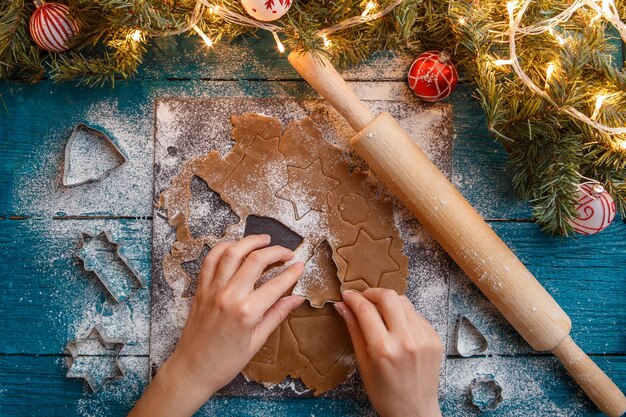 Foto de mãos humanas, rolo, massa, ramos de abeto, guirlandas, moldes de biscoito na mesa azul