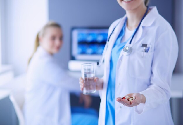 Foto de médico segurando pílulas e copo de água na clínica com colega no fundo