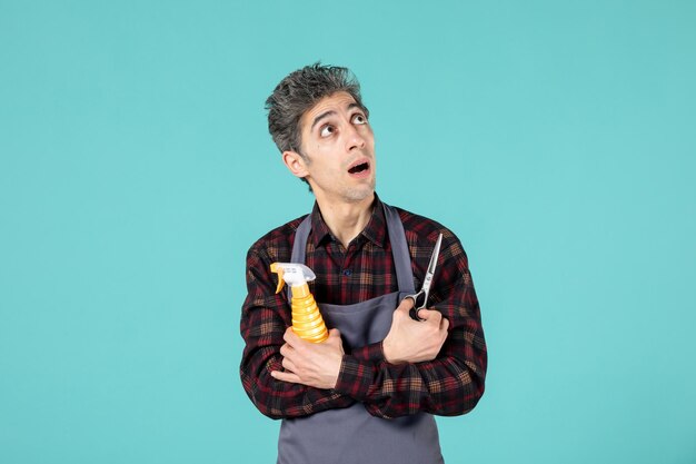 Foto de meio corpo de cabeleireiro chocado usando avental cinza e segurando um frasco de spray de tesoura sobre fundo de cor azul pastel