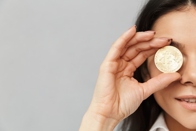 Foto de meio lado de mulher rica com longos cabelos castanhos cobrindo os olhos com bitcoin dourado, isolada sobre parede cinza