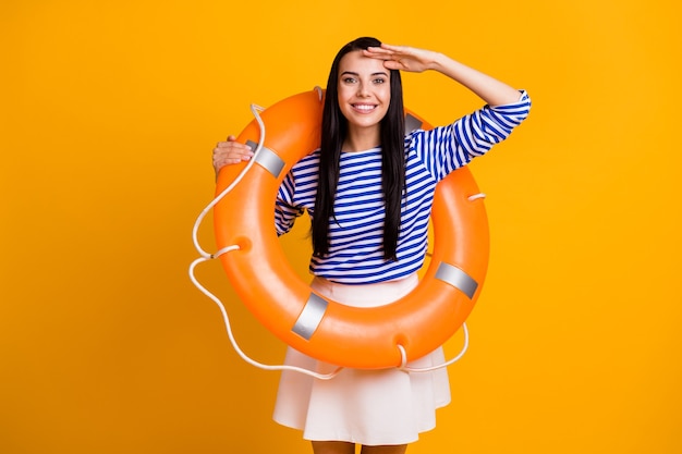 Foto de menina alegre positiva com bóia salva-vidas segurar mão rosto ver terra do oceano água usar camisa listrada saia vestido branco azul isolado sobre fundo de cor de brilho brilhante