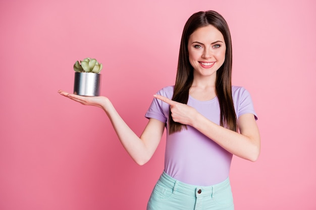 Foto de menina positiva segurar vaso de flor ponto dedo indicador presente ecologia objeto de segurança usar calça azul-turquesa azul-petróleo isolado sobre fundo de cor rosa