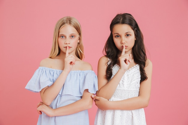 Foto de meninas sérias de 8 a 10 anos de idade usando vestidos segurando o dedo indicador nos lábios e pedindo para manter silêncio, isolada sobre um fundo rosa