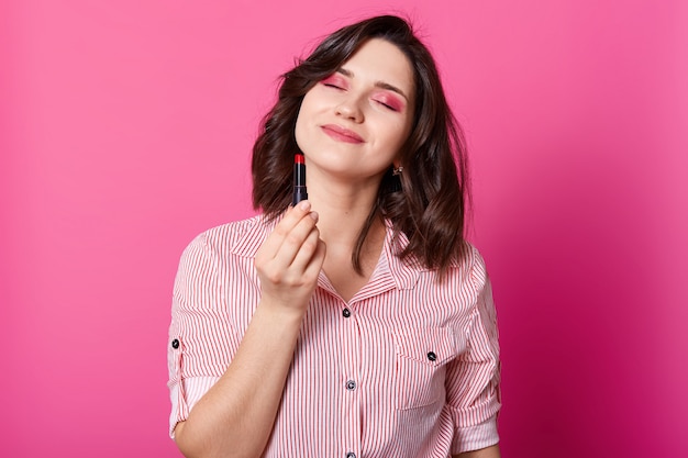 Foto de metade do comprimento da mulher fica com os olhos fechados e segurando o batom vermelho.