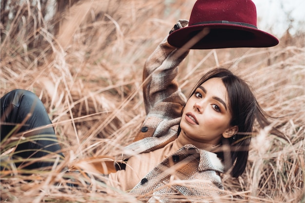 Foto de moda ao ar livre da bela jovem em paisagem de outono com grama seca e chapéu