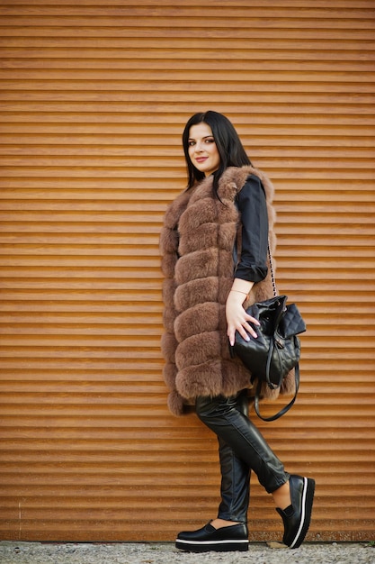 Foto de moda ao ar livre de linda mulher sensual com cabelos escuros em roupas elegantes e casaco de pele luxuoso e com mochila contra o fundo do obturador