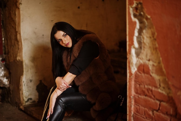 Foto de moda ao ar livre de linda mulher sensual com cabelos escuros em roupas elegantes e casaco de pele luxuoso na rua velha com paredes de grunge