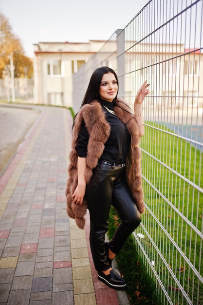 Foto de moda ao ar livre de linda mulher sensual com cabelos escuros em roupas elegantes e luxuoso casaco de pele sem mangas contra cerca de ferro na cidade de outono