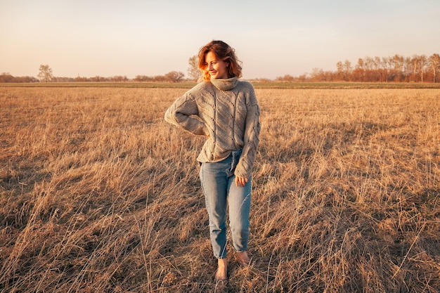 Foto de moda ao ar livre de uma jovem bonita em suéter de tricô marrom e jeans na paisagem de campo de outono Lookbook de moda