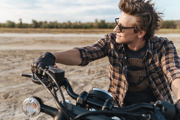 Foto de motociclista jovem positivo bonito em bicicleta ao ar livre no campo deserto, olhando de lado.