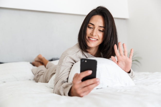 Foto de mulher adulta de 30 anos usando telefone celular, deitada na cama com lençóis brancos em um quarto bem iluminado