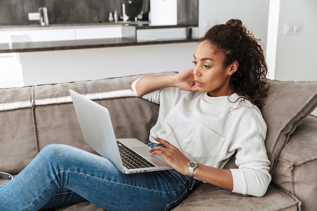 Foto de mulher afro-americana satisfeita usando laptop, enquanto estava deitado no sofá na sala de estar brilhante