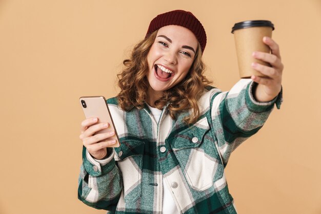 Foto de mulher alegre com chapéu de tricô usando celular e bebendo café para viagem isolada em bege