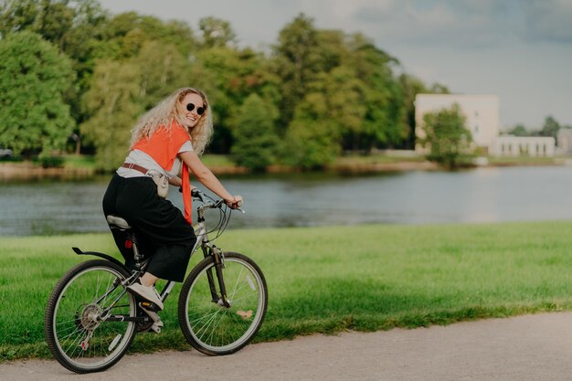 Foto de mulher alegre vestida casualmente, anda de bicicleta, olha de lado, tem expressão feliz, usa óculos escuros, posa perto de rio, gramado verde e árvores, alguns prédios ao fundo. Gente e descanso