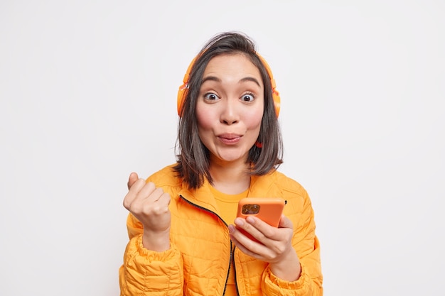 Foto de mulher asiática bonita com cabelo escuro cerrando o punho usa smartphone para comunicação online ganha nova assinatura de música vestida com jaqueta laranja isolada sobre parede branca