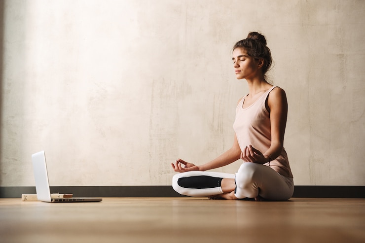 Foto foto de mulher atraente concentrada em roupas esportivas meditando com os dedos zen e usando o laptop enquanto está sentada no chão em casa